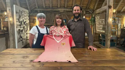 Sara Johnson, with brown bobbed hair and wearing a red and white blouse, holds her late father's faded red Manchester United shirt from 1958. To her right, Rebecca Bissonnet has short grey hair and wears black dungarees over a white t-shirt. To Sara's left, Dominic has dark hair and a beard. He is wearing an open-collared dark grey shirt.