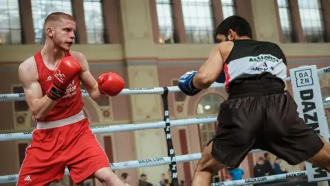 Handout Bobby is on the left of the picture, wearing a red boxing vest and shorts, along with red gloves. He is facing a boxer who is dressed in a black top and black shorts.