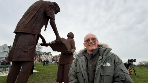 JIM SCOTT/BBC David Gibson, who is Len Gibson's son, is wearing a light green North Face jacket and is standing in front of the artwork of his father. He is wearing brown tinted glasses and is smiling. In the background there are some people watching after the unveiling has taken place.