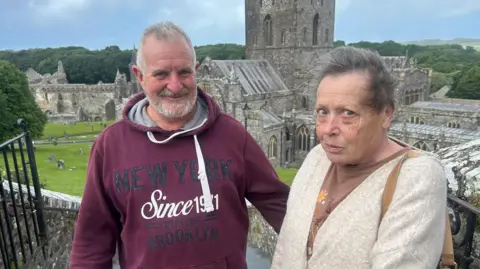 Paul Collins and Lin Palfrey standing outside the Cathedral. Paul, on the left, wears a burgundy hoodie and has short grey hair. Lin stands next to him and wears a beige cardigan and brown t-shirt. She has short grey hair. 