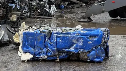 A block of crushed blue, black and white metal on a wet road surface with pieces of metal in the background
