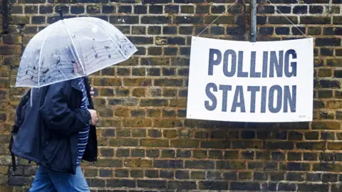 A person holding an umbrella walks past a polling station sign