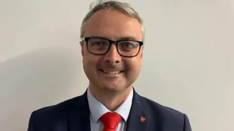 Juan Watterson mirando directamente a la cámara, sonriendo.  Lleva una chaqueta azul marino, camisa azul pálido y corbata roja brillante, junto con gafas de montura negra.  Tiene un pin rojo con un símbolo amarillo de las tres patas de Mann en el ojal de su chaqueta.