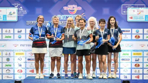 Pamela Peard Eight competitors all stand joyfully on a blue podium, adorned with medals and holding small blue plushie whales. Front and centre is Sian and Pam, wearing matching pale green-blue t-shirts with navy stripes across them. Sian is wearing a blue skirt and trainers, whilst Pam is wearing black shorts and white trainers. On the wall behind them are multiple sponsors for the event, with 'amazing Thailand' written along the top.
