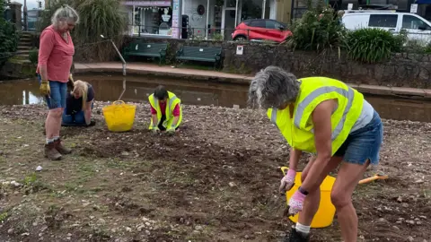 Four female volunteers work to pull weeds from the soil by a brook.