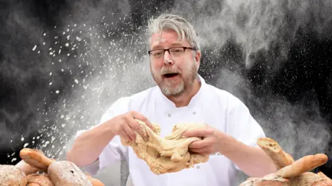 Chris Taylor Richard Hughes in chef's whites, kneading dough and surrounded by loaves