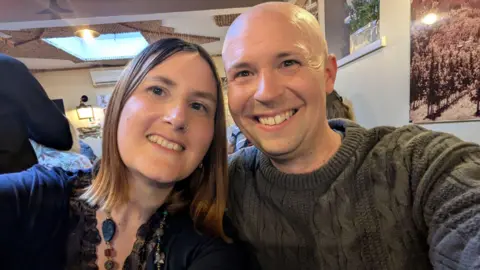 Becky and Uan in a selfie together. Both are smiling from a room that has other people in it. Beck has long dark hair and wears a blue t-shirt and necklace. Uan is bald and wears a thick grey cable knit jumper.