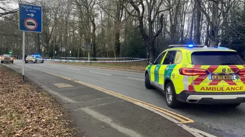 A carriageway with a large police cordon sectioning off a length of the road. There are police cars ahead an ambulance car directly at the front of the image