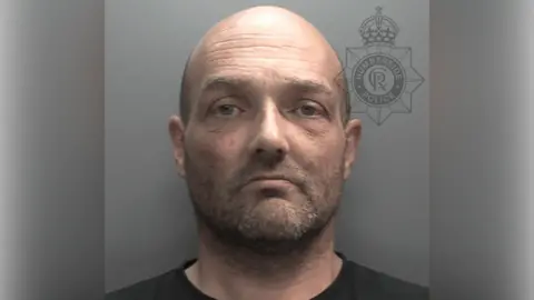A police custody shot of Alan Wigglesworth. He is bald, with facial stubble, and is wearing a black t-shirt. The wall behind him is grey.
