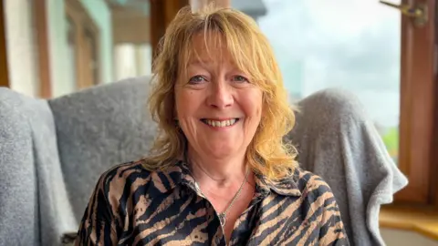 A woman smiling for the camera. She sits on a chair with a grey blanket over it. She has blonde hair and is wearing a tiger-print top