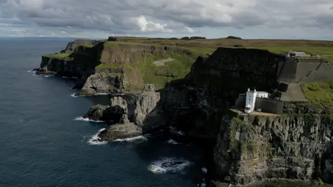 A dramatic view of cliffs on Rathlin Island with a calm sea in the foreground. A white building called Rathlin West Lighthouse is perched half way up one of the cliff on the right hand side of the photo.