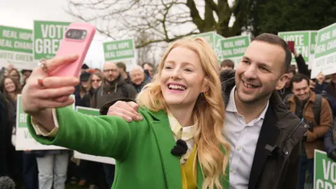 Hannah Spencer and Zack Polanski pose together as she takes a selfie with her pink mobile phone. A crowd of people stand behind in a park holding Vote Green placards