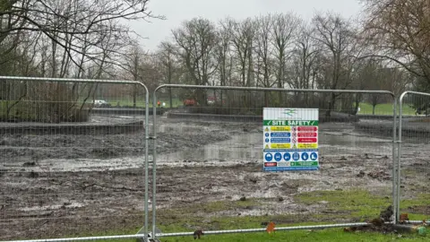 Image shows a lot of sludge and mud where the water would have been in the lake. There is some water but very little and there is a mound with trees on in the middle of where the lake was. In front of the lake area are metal fences and a sign which says site safety on it and warning signs alerting people not to enter the area. 