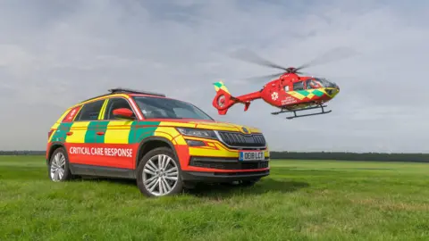 A car and a helicopter belonging to Thames Valley Air Ambulance photographed on a cloudy day. The parked car has a sign on the side that reads "Critical Care Response". The helicopter is in the air.