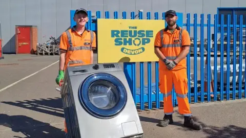 Two men in orange high visibility clothing. There is a blue fence behind them with a yellow sign that says "The reuse shop Devon". The man on the left is carrying a silver washing machine. The 