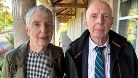 Brother and sister, Ellen and Jimmy Dorris. Ellen, on the left, wears a black and white top and green jacket. She has short grey hair. Jimmy wears a striped shirt and diagonally striped tie. He is also wearing a black coat. 