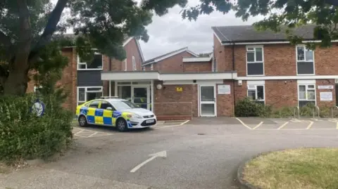 Ben Parket/BBC Police car parked outside a red-brick, double-storey housing complex. The is a car park to the front of the building and a green patch of grass. 