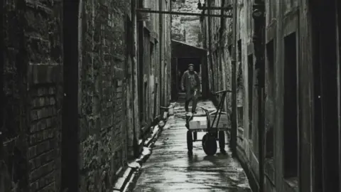 A black and white photo of a narrow passageway with a workman and a cart 