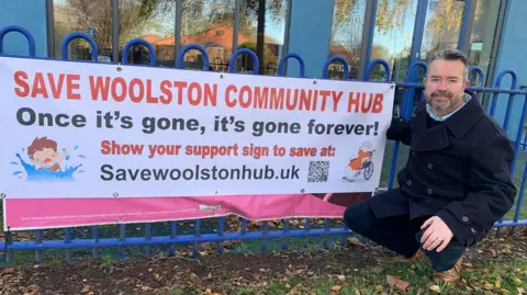 Mark Evans-Guillen crouches in front of banner saying "Save Woolston Community Hub - Once it's gone, it's gone forever". He is in front of railings in front of the site. 