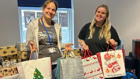 ARC A photo of two women holding Christmas bags with wrapped parcels piled up on a table in the background