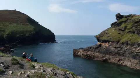 Boscastle harbour on the South West Coast Path. There is a man and a woman sitting on the steep slope leading to an inlet with another rocky outcrop on the other side. To the left is a headland. It is a clear day.