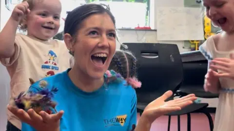 The Way Youth Zone A woman with tied-back dark hair and a blue jumper which says "The Way" laughs as she sits on the floor while two young girls put feathers on her shoulders and hands.