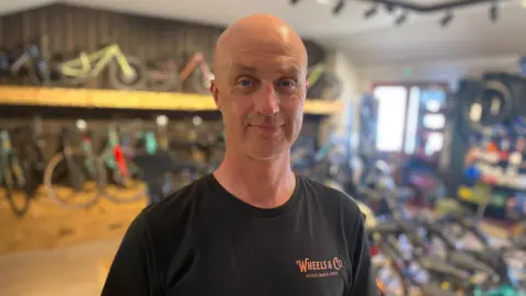 Matthew stands in a bicycle shop, wearing a black Wheels and Co. shirt, with various bikes displayed behind on racks and floor, under warm lighting and blurred indoor background.