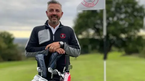 Mark Butler Mark está vestindo um top e calças de golfe e está atrás de um conjunto de tacos. Ele tem cabelos grisalhos e barba.