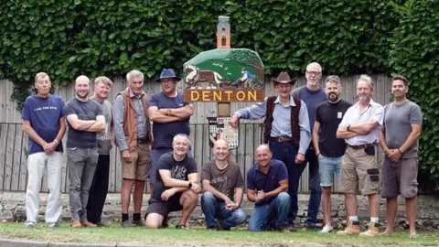 Mark Richards A group of men pose before a village sign. they are all smiling