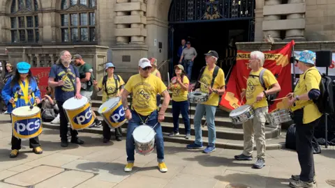 A group of people are wearing yellow T-shirts which say "PCS" and are playing hand-held drums on the steps of Sheffield Town Hall.
