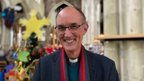 Reverend Kelvin Inglis in front of decorated Christmas trees in a church. He is wearing spectacles, a dark jacket, red scarf and green sweater