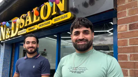 Two men with neat dark beards smile outside a shop front.