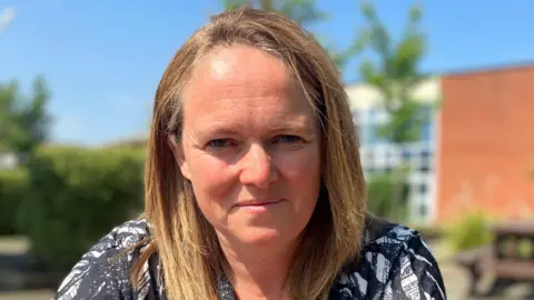 A women with light brown hair is looking directly at the camera. She is wearing a black and white patterned top. She is in an outdoor location with blue sky and some plants in the background.