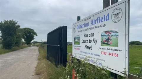 The entrance to Otherton Airfield showing an open black gate with a road going past it. A white sign just before the gate on the right says "Otherton Airfield" in blue letters with contact details, slogans and two pictures of aircraft. 