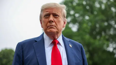 Getty Images Donald Trump has light coloured hair stares off camera. He has a neutral expression. He wears a blue suit, light blue shirt and red tie. On his lapel is a USA flag pin