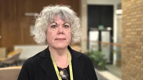 Paul Moseley/BBC A picture of a woman with curly grey hair standing indoors, wearing a dark jacket and a lanyard. The background shows a modern public building with wood-panelled walls, brickwork, seating areas and soft, natural lighting.
