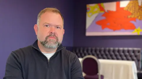 Andrew sits in a purple-walled room on a bench-style seat with colourful pop‑art style paintings behind them. He is wearing a dark jumper and have their hands clasped in front of them.