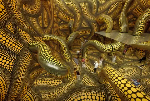 AFP via Getty Images A series of intertwining tentacles in black and yellow polka dot run through a large room with mirrors in the centre, with visitors walking through it, in Basel in 2025.