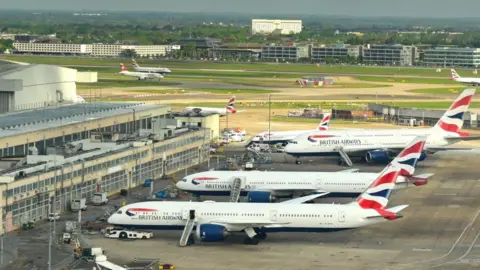 Planes at Heathrow Airport. Three British Airways planes are prominent in the photo, facing a yellow building, which is on the left. Other planes, grass and buildings are in the distance.