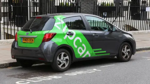 Getty Images A small grey hatchback car is parked on the street in front of a row of terraced houses. It has bright green Zip Car branding on one side.