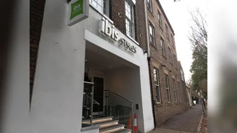 An exterior shot of Ibis Style Hotel in York, and red-brick building with a white plastered facade around the entrance, and the green Ibis style logo sign above. In the entrance, a short flight of stairs can be seen, alongside an accessible lift which has two orange cones placed in front of it. 