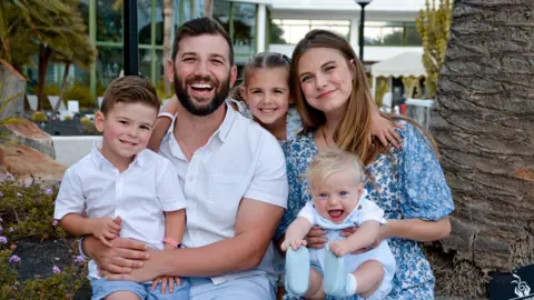 Hannah Moore A man with dark hair and beard in a white shirt sat next to a woman in a blue and white floral dress. The man is holding a young boy on his lap also in a white shirt. The woman is holding a baby in a white shirt on her lap. Between the adults is a young girl's head. They are sat next to a tree trunk