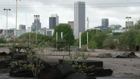 Wasteland stands in the foreground with the city centre in the distance. Tyres are lying on the ground. Tall buildings are seen in the skyline.