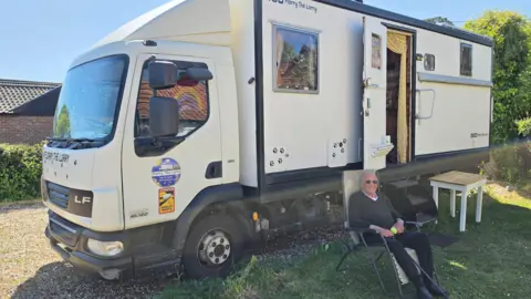Supplied An older man wearing a grey jumper and black trousers holding a walking stick while sitting in a chair in front of a large white lorry.