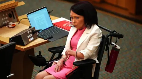 PA Media Pam Duncan-Glancy, who has short dark hair, sits in a wheelchair at a desk in the Scottish Parliament. Sitting side on, she is wearing a white jacket over a pink dress. There is an open laptop on her desk.
