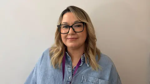 A woman with blonde hair and black framed glasses. She's wearing a denim blue top.