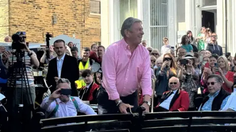 Alan Titchmarsh holds scissors as he cuts a ribbon to open the Ilkley Fountain, surrounded by a crowd.