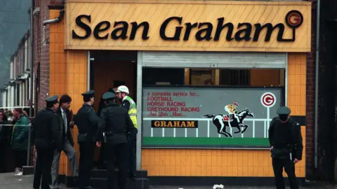 Pacemaker Archive image of Sean Graham bookmakers shooting. The front of the building is yellow with black writing of 'Sean Graham'. A group of police officers are outside.