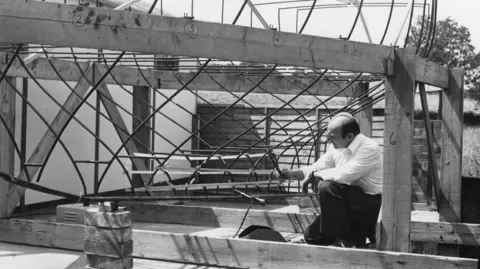 Phil Boocock A black and white image of Lee Holmes wearing a shirt and trousers and looking at a partial frame of a boat