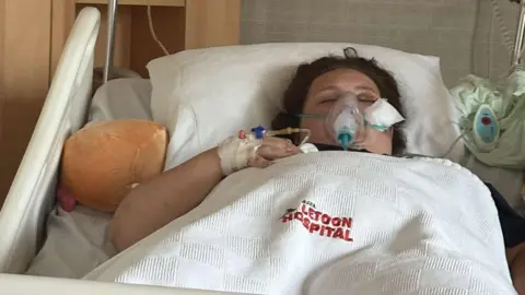 Meggan Nichol Lily is lying in bed in hospital. There is a breathing mask over her face and her hand is in a bandage with an IV drip in it. You can see hospital equipment like an IV in the background